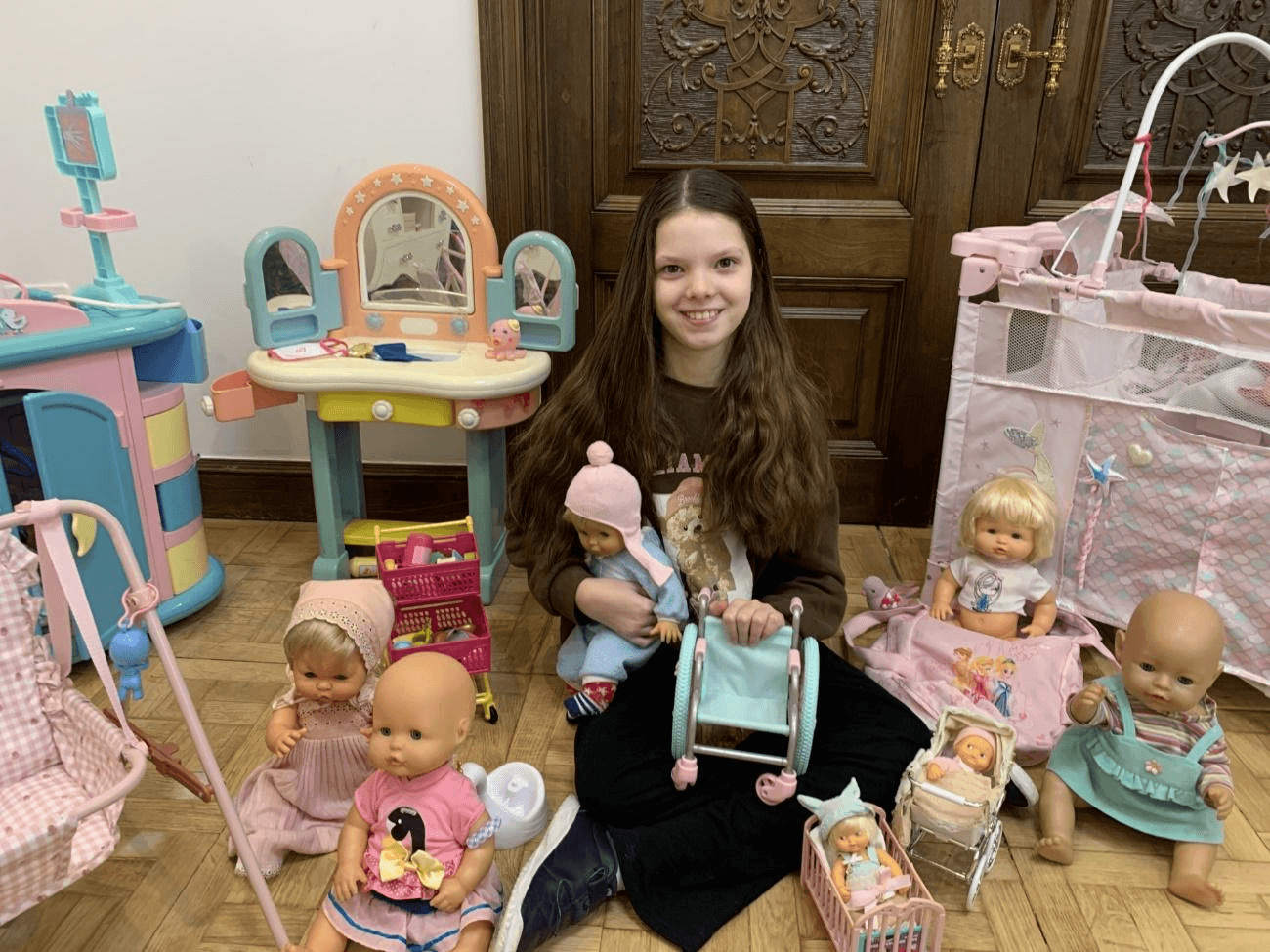 Una niña sonríe sentada en el suelo, rodeada de muñecas y juguetes, sosteniendo una silla de ruedas de juguete.