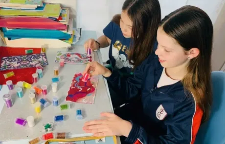 Two COAMI pupils apply coloured glitter to their drawings in class.
