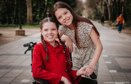 Two sisters smile in a park; the elder one, in a red coat, is in a wheelchair.