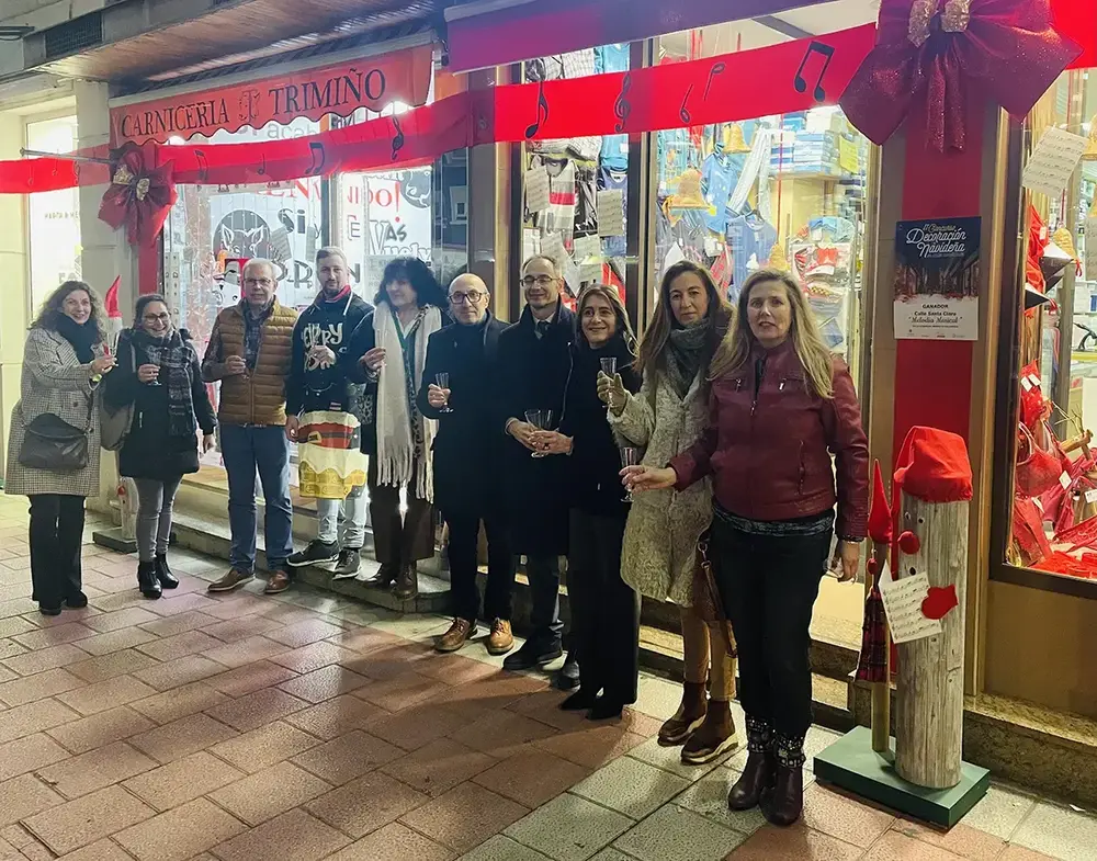 A group raises their glasses outside a shop decked with red ribbons and musical notes, beside the Christmas contest sign.