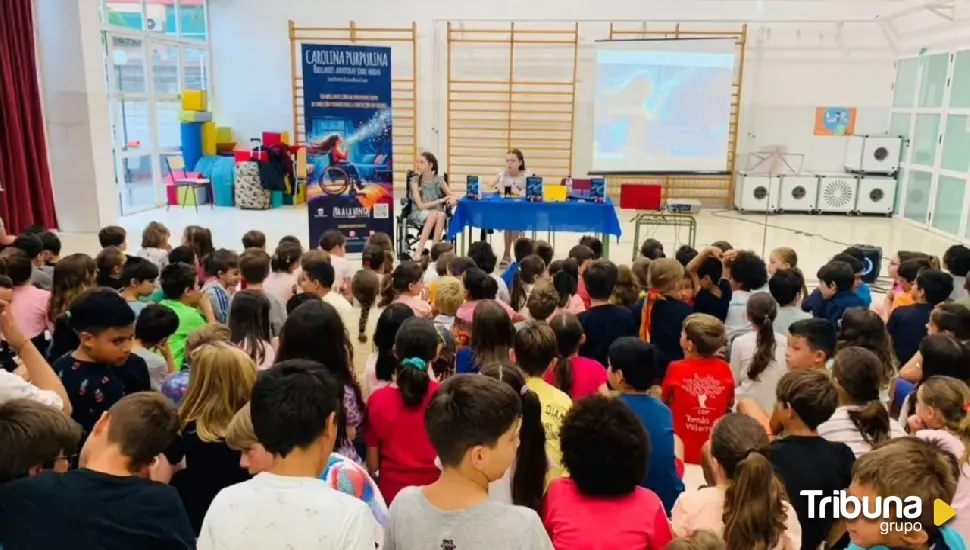 Dozens of schoolchildren sit on the floor listening to two girls, one in a wheelchair, beside the book’s banner.