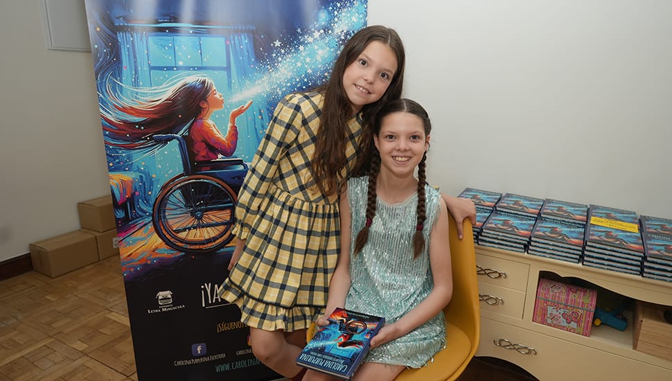 Two girls pose beside the book’s banner; one holds a copy, with stacks of books behind them.