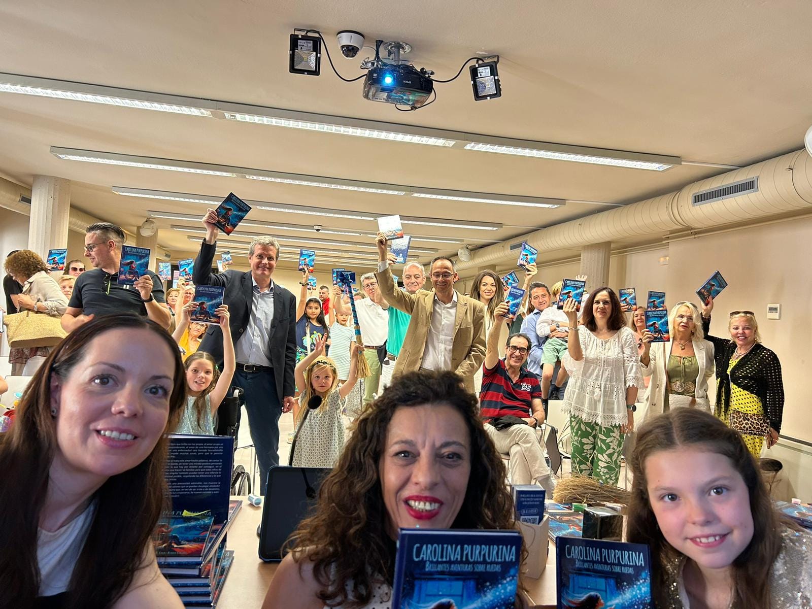 Selfie at the launch: the audience holds up copies of “Carolina Purpurina” above a table stacked with books.