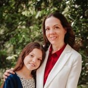 Outdoor portrait: a woman in a white jacket with her arm around a smiling girl, against leafy greenery.