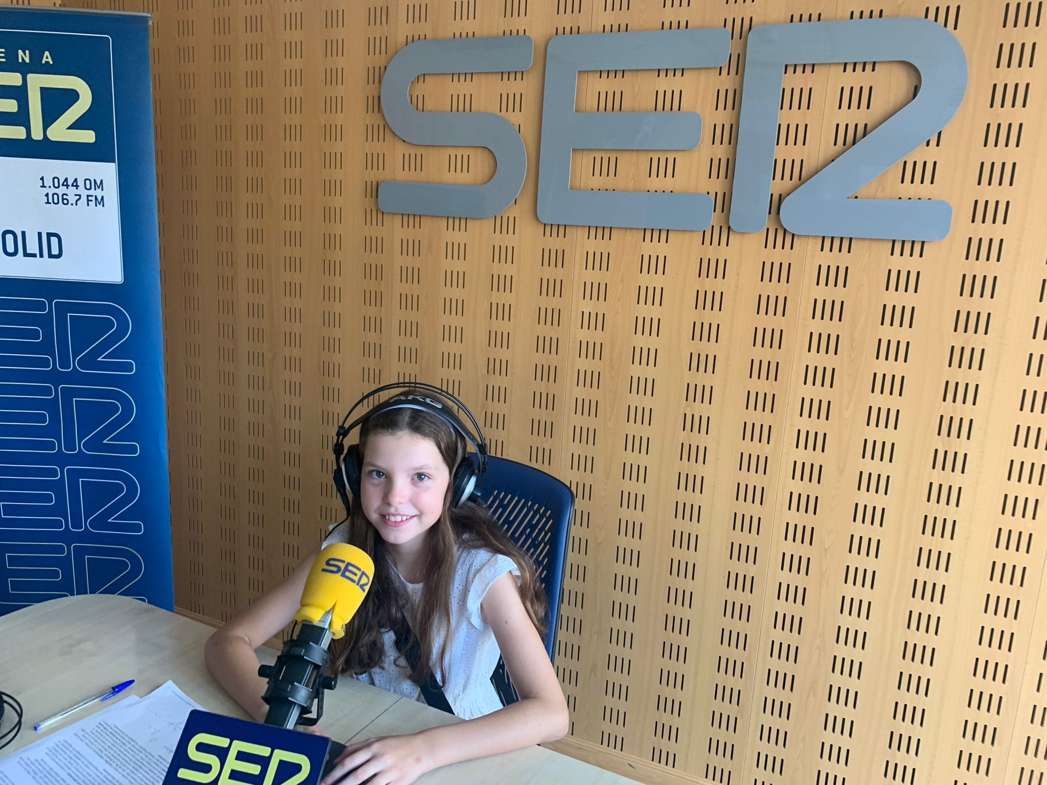 A girl in headphones smiles at the yellow Cadena SER microphone, under the station’s logo in the studio.