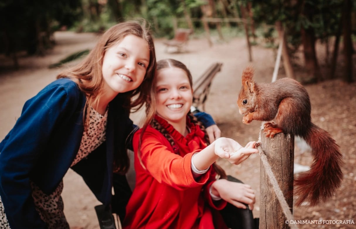 Two girls smiling in a park; the one in a wheelchair offers food from her palm to a squirrel perched on a post.