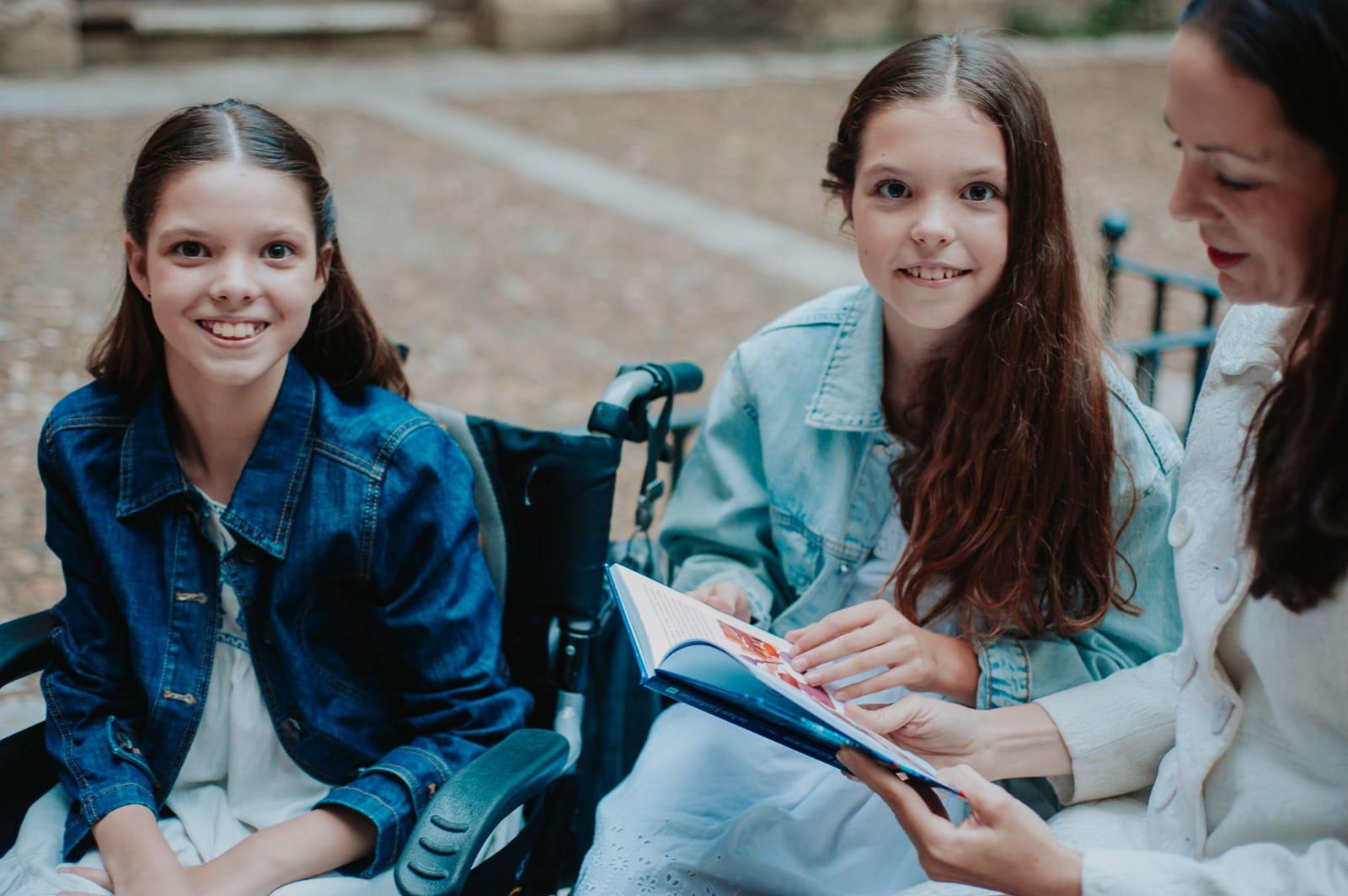 Outdoors, a girl in a wheelchair beside another girl and a woman leafing through the open book.