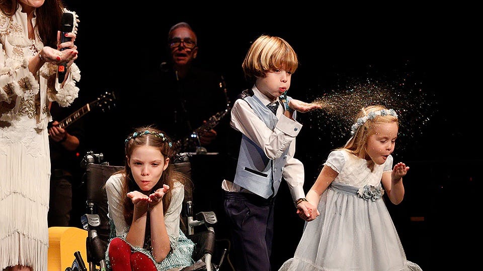 On stage, three children blow glitter; one of them, in a wheelchair, wears a flower crown.