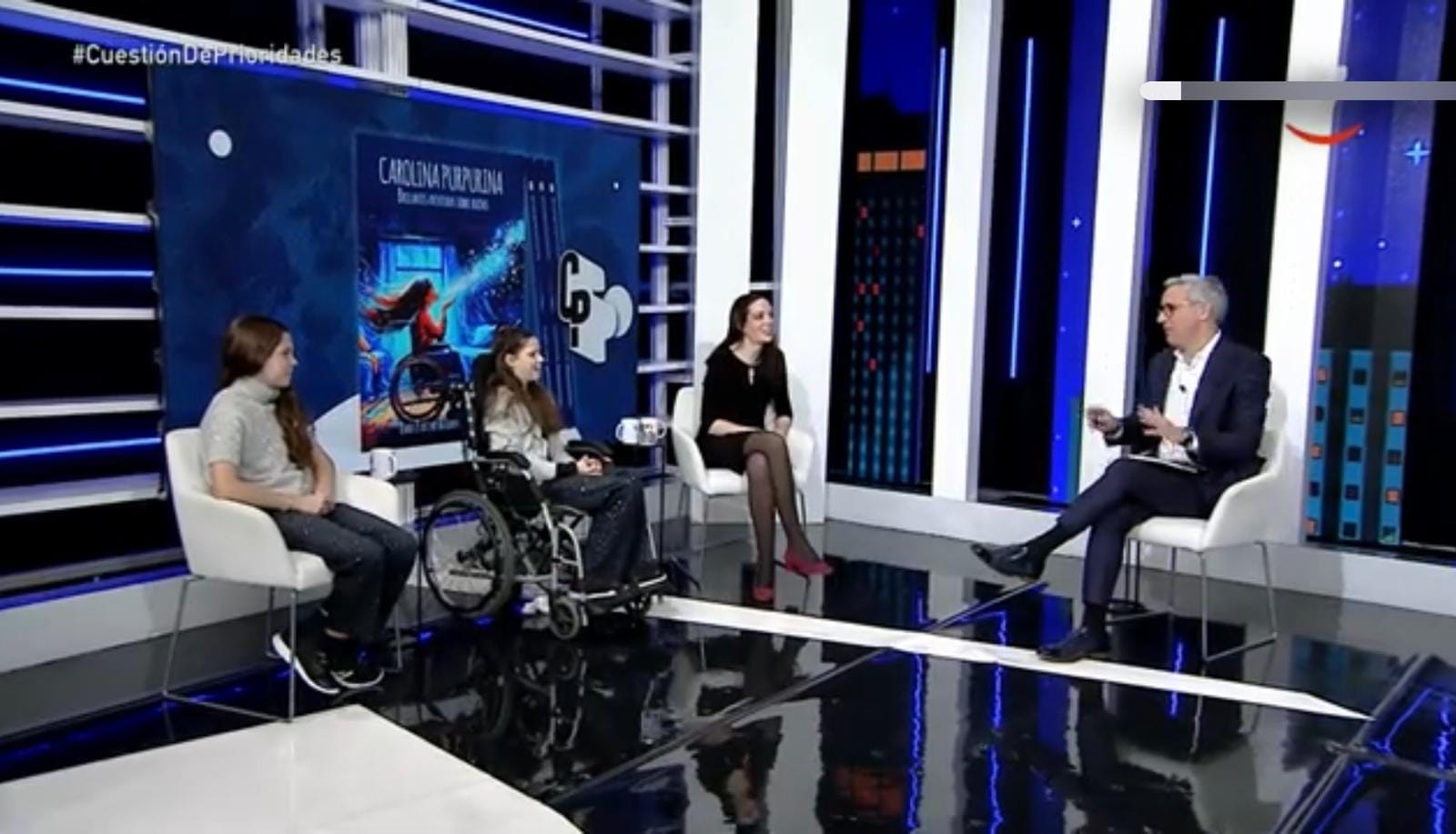 Television studio: two girls, one in a wheelchair, and a woman talking with the presenter beside the book cover on screen.