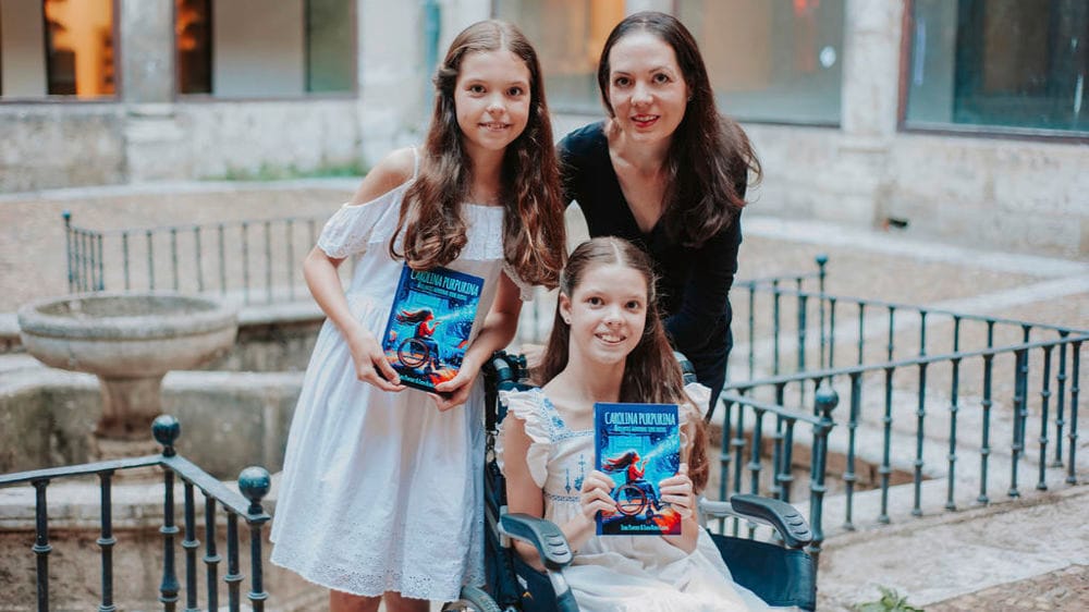 Two girls hold a copy of “Carolina Purpurina” in a courtyard; one uses a wheelchair, and a woman stands behind them.