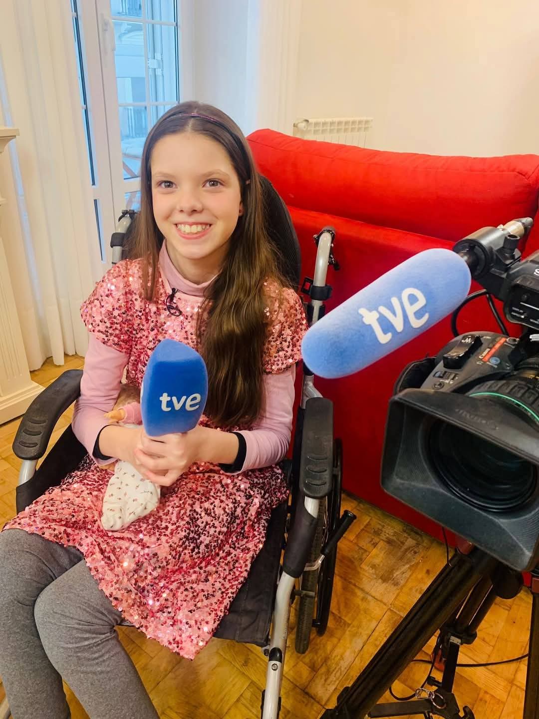 Carolina, in her wheelchair and a pink sequinned dress, holds a TVE microphone in front of the camera.