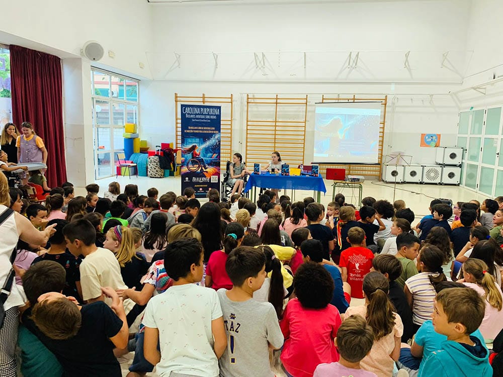 Dozens of schoolchildren sit on the floor listening to two girls at the table, one in a wheelchair, beside the banner.