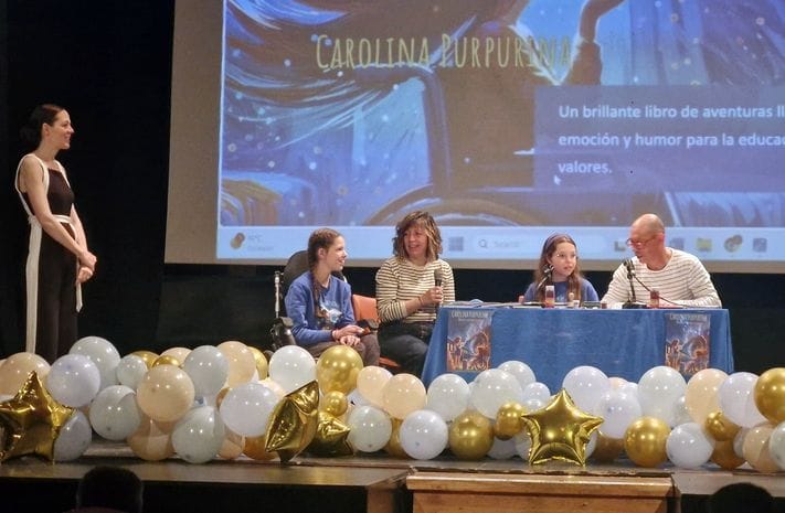 A talk on a school stage: four people at the table, one at the microphone, among gold and white balloons.