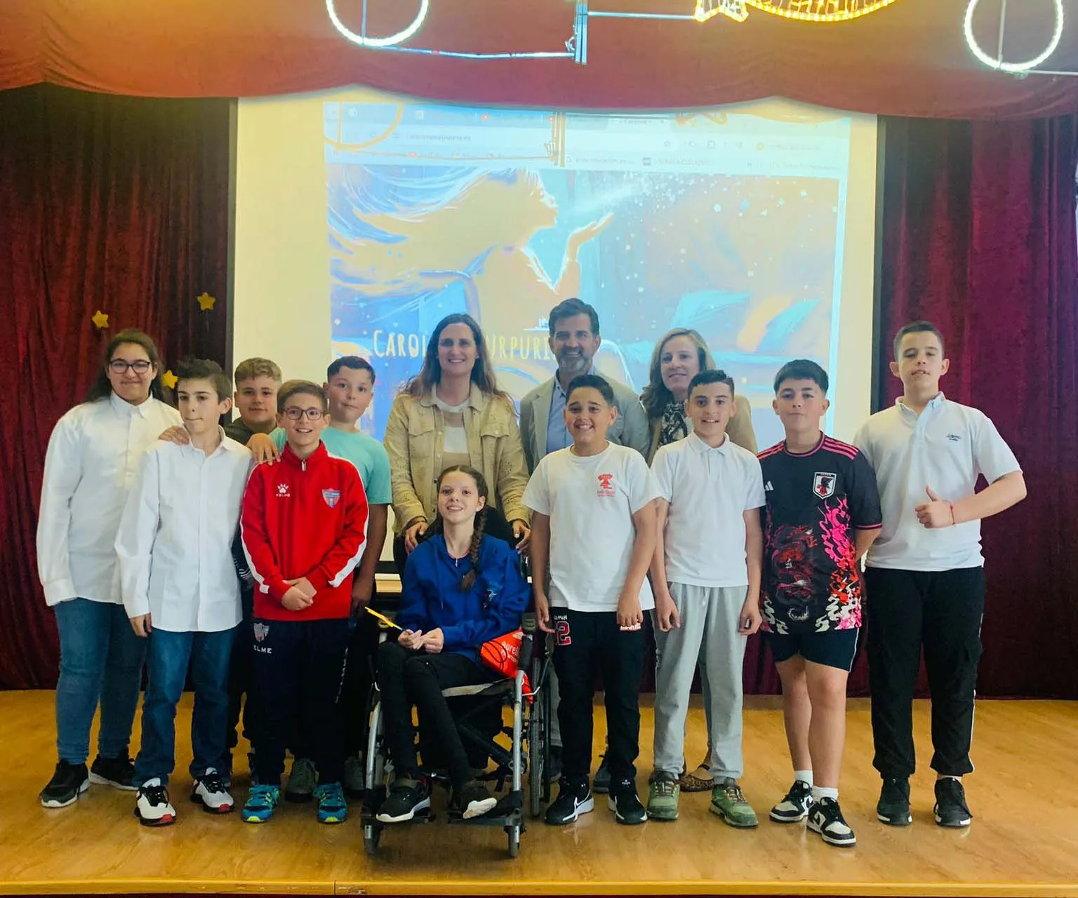 Group photo on stage: pupils gather around Carolina in her wheelchair, with the book cover projected behind them.