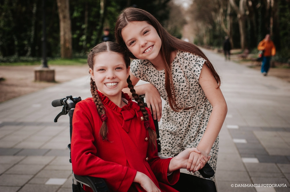 Carolina, in her wheelchair and red coat, smiles beside another girl hugging her on a tree-lined walkway.