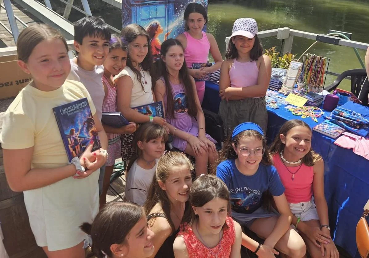 A group of girls poses beside the riverside book stall; several of them are holding the book “Carolina Purpurina”.