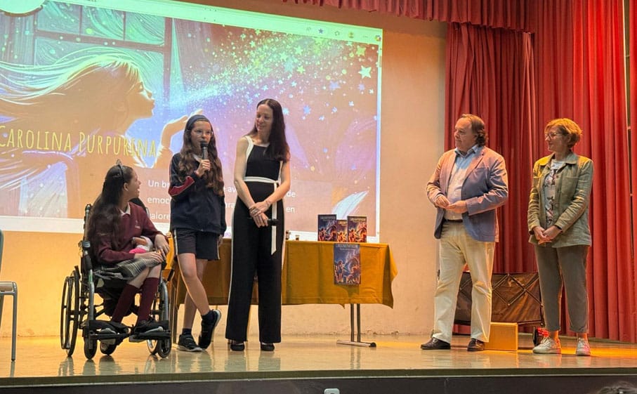 On stage, a teenager speaks into the microphone beside Carolina in her wheelchair, the book cover projected behind them.