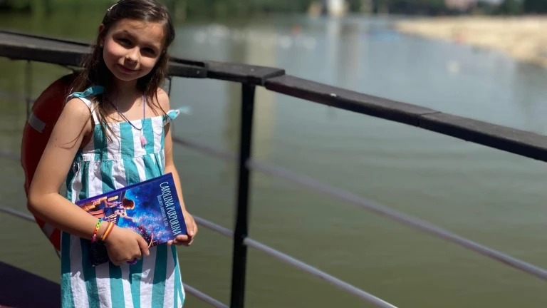 A girl in a striped dress with a backpack holds a copy of “Carolina Purpurina” by a railing beside a river.