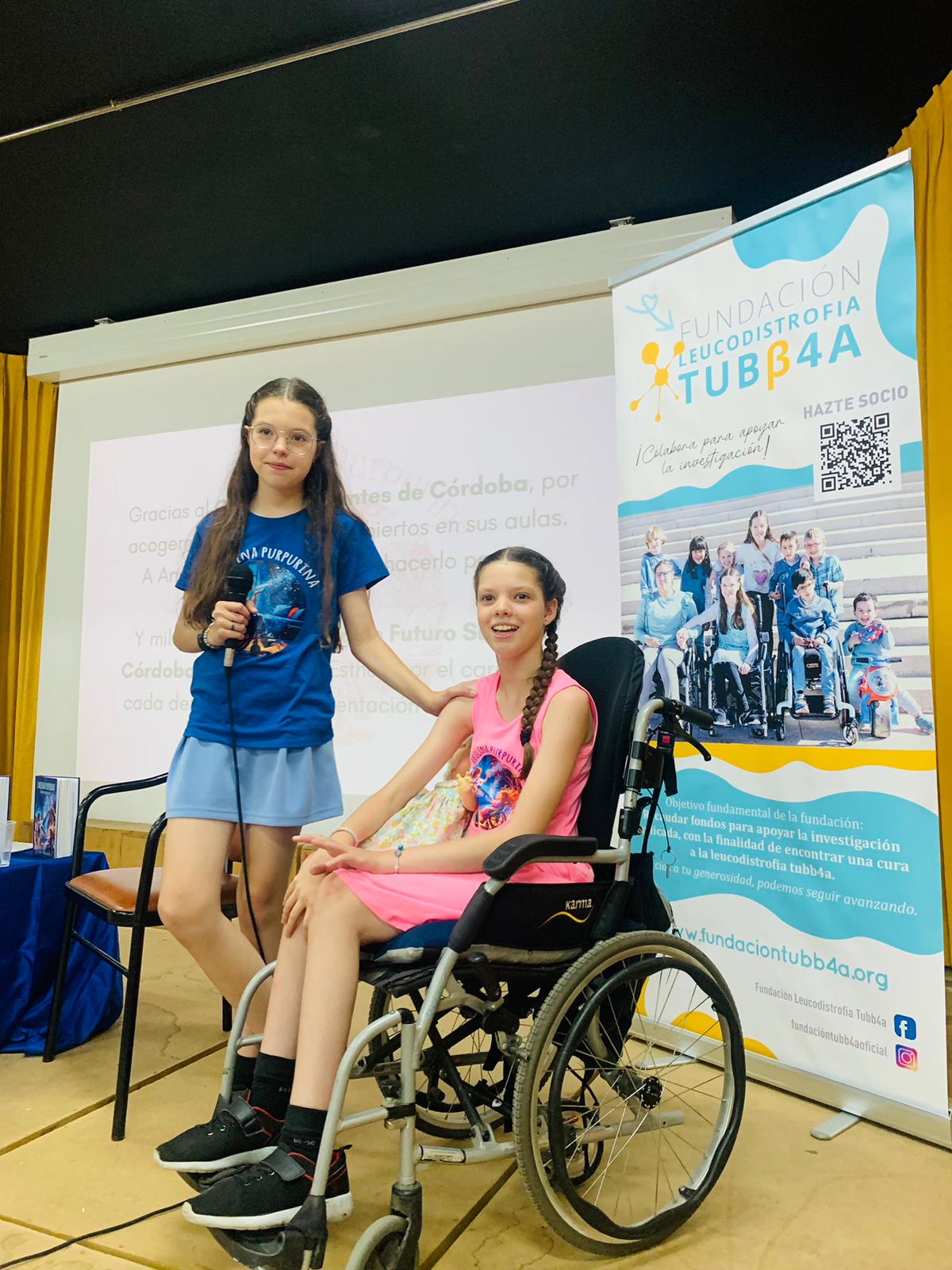 Isabel, holding a microphone, beside Carolina in her wheelchair, in front of the Fundación Leucodistrofia TUBB4A banner.