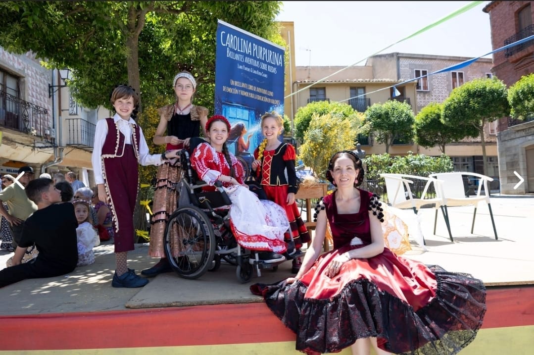 On a stage, children in period costume beside the book’s banner; one is in a wheelchair, another blows a handful of glitter.
