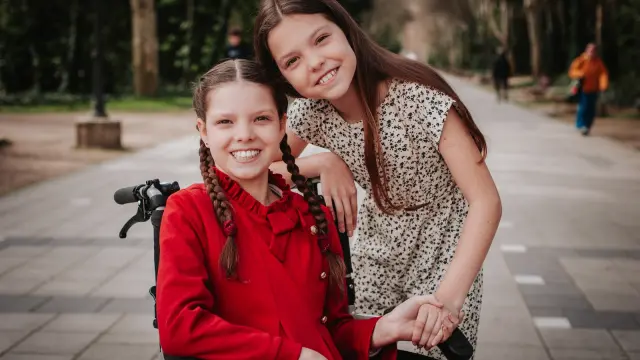 Carolina smiles in her wheelchair while another girl leans on her shoulder along a tree-lined path.