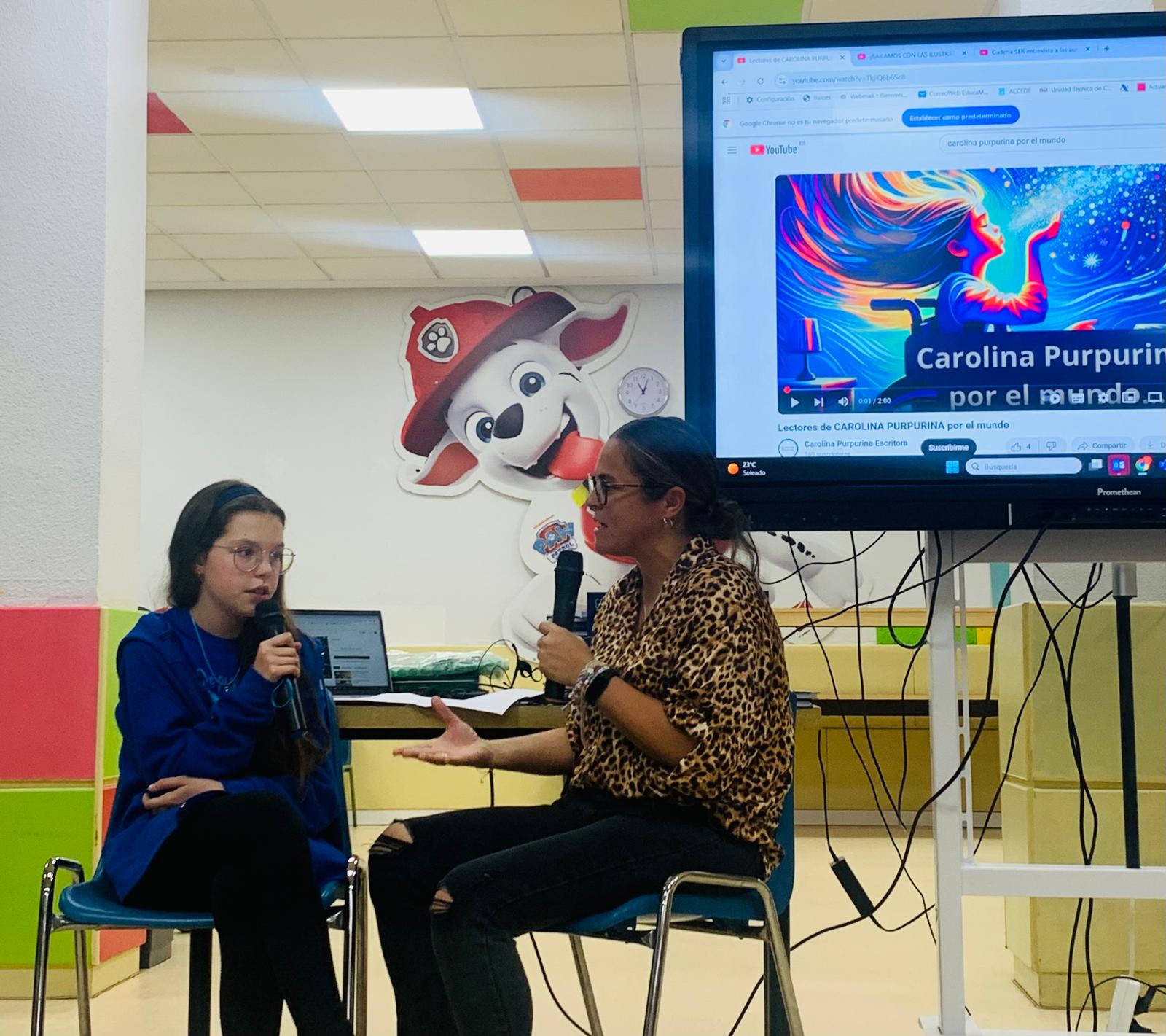 Isabel, microphone in hand, answers an interviewer in front of a screen playing the video “Carolina Purpurina por el mundo”.