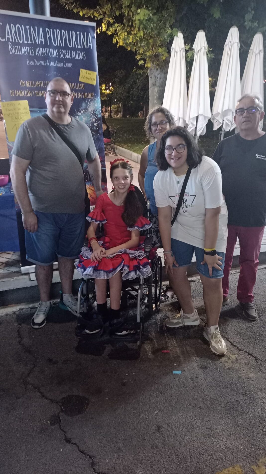 At the fair by night, a girl in a flamenco dress in her wheelchair poses with several people in front of the book’s banner.