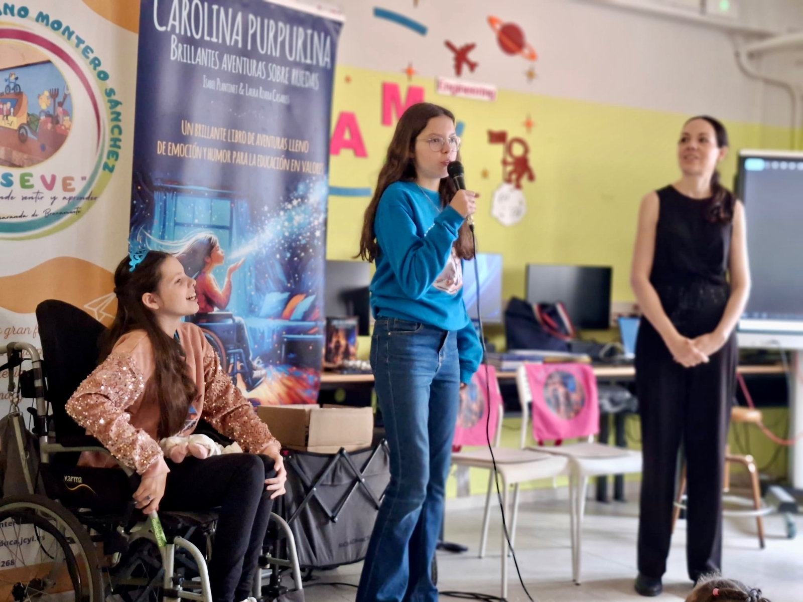Isabel speaks into the microphone during the school visit, with Carolina and Laura beside her.
