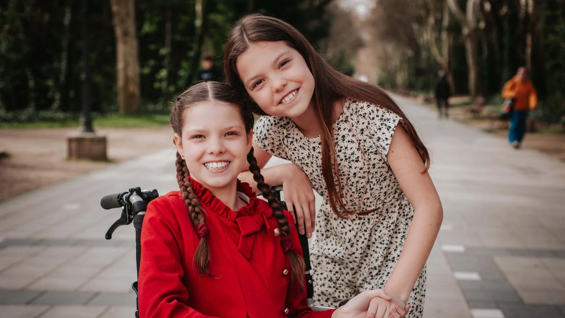 Carolina, in a red coat and braids, smiles in her wheelchair beside another girl holding her hand, on a tree-lined path.