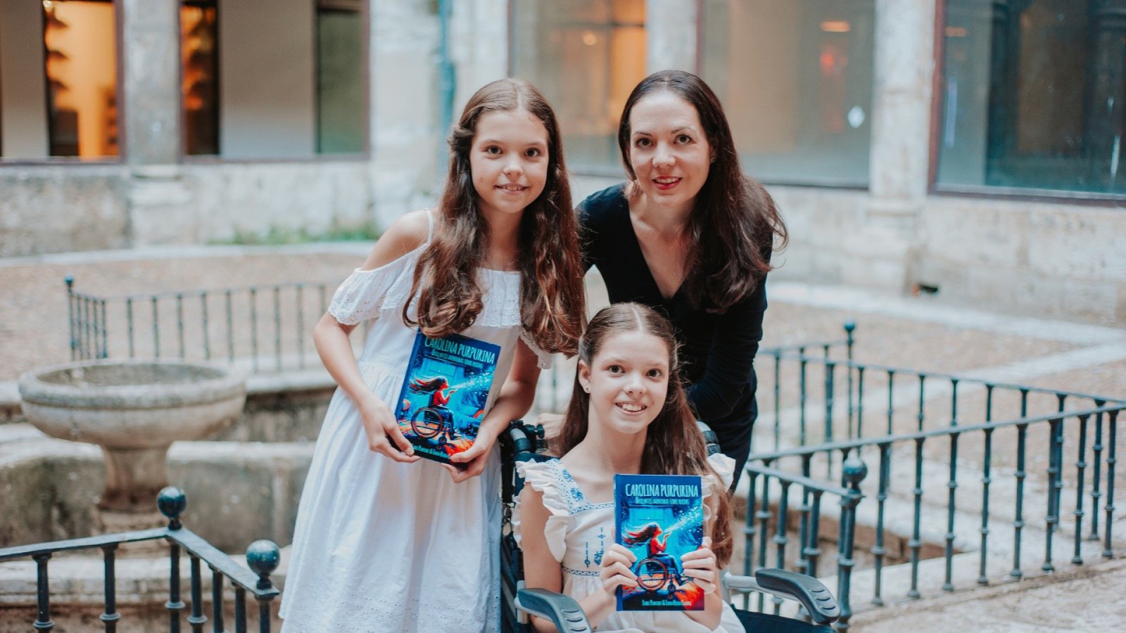 Carolina, in her wheelchair, and another girl hold up the book “Carolina Purpurina” next to a woman in a stone courtyard.