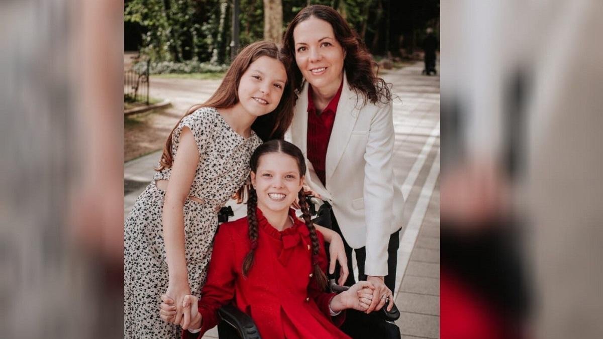 Carolina, in a red dress with braids, smiles in her wheelchair beside a girl in a patterned dress and a woman in a white jacket.
