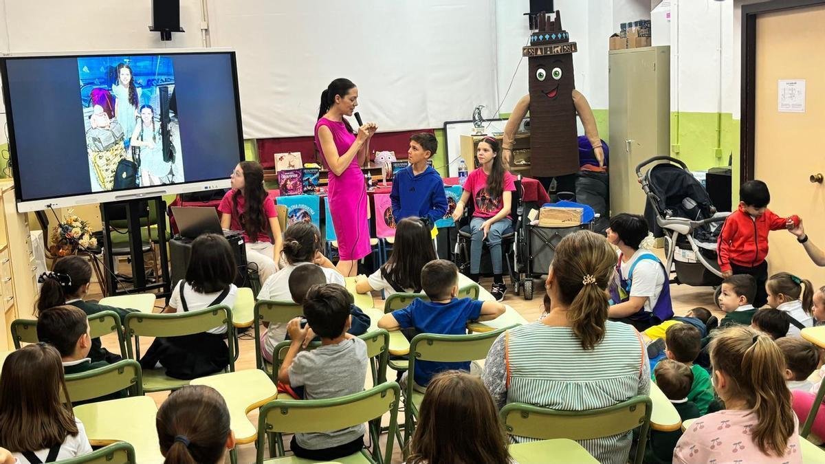 A woman speaks into a microphone to seated pupils; beside her are several children and Carolina, in her wheelchair, by a table of books.