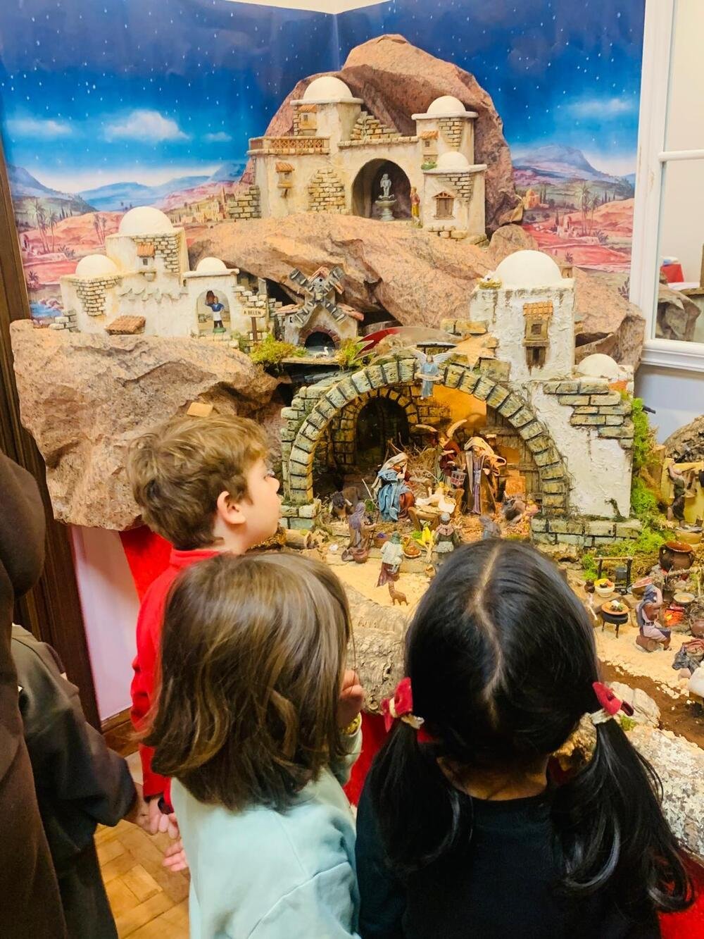 Three children, seen from behind, look at the nativity scene: white houses, a stone stable with the Nativity and a starry sky backdrop.