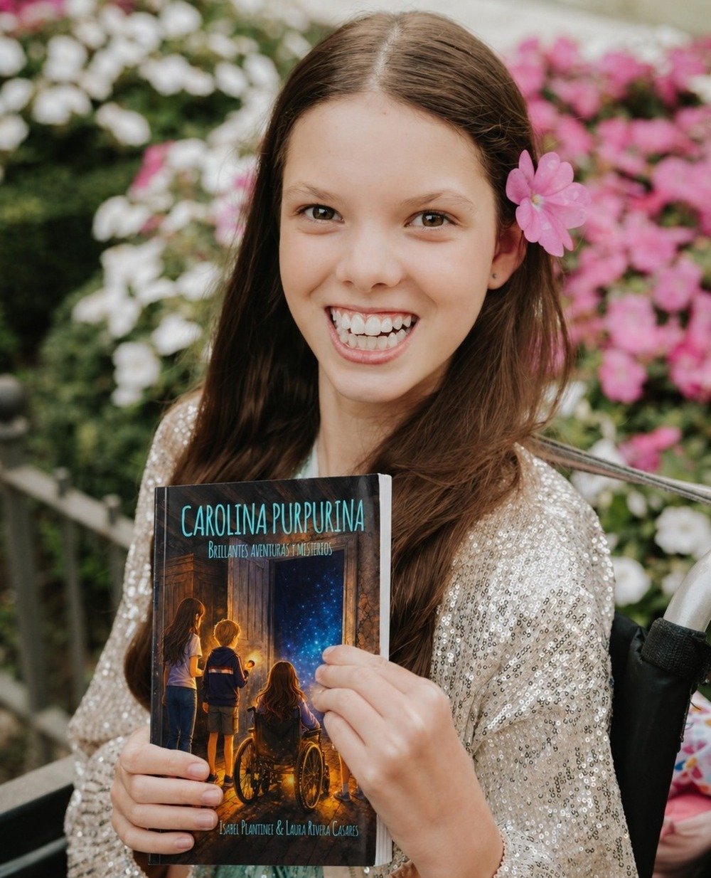 A smiling girl with a pink flower in her hair holds the book “Carolina Purpurina. Brillantes aventuras y misterios”.