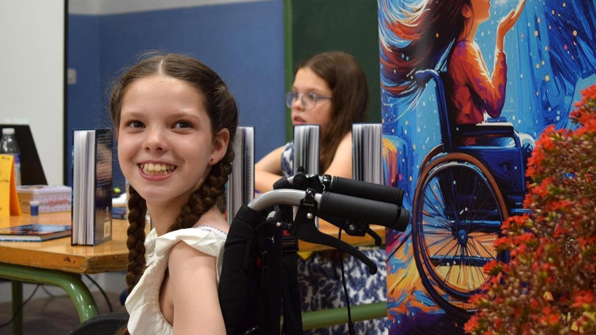 Book presentation: Carolina smiles in her wheelchair beside the cover banner; behind her, a girl sits at the table of books.
