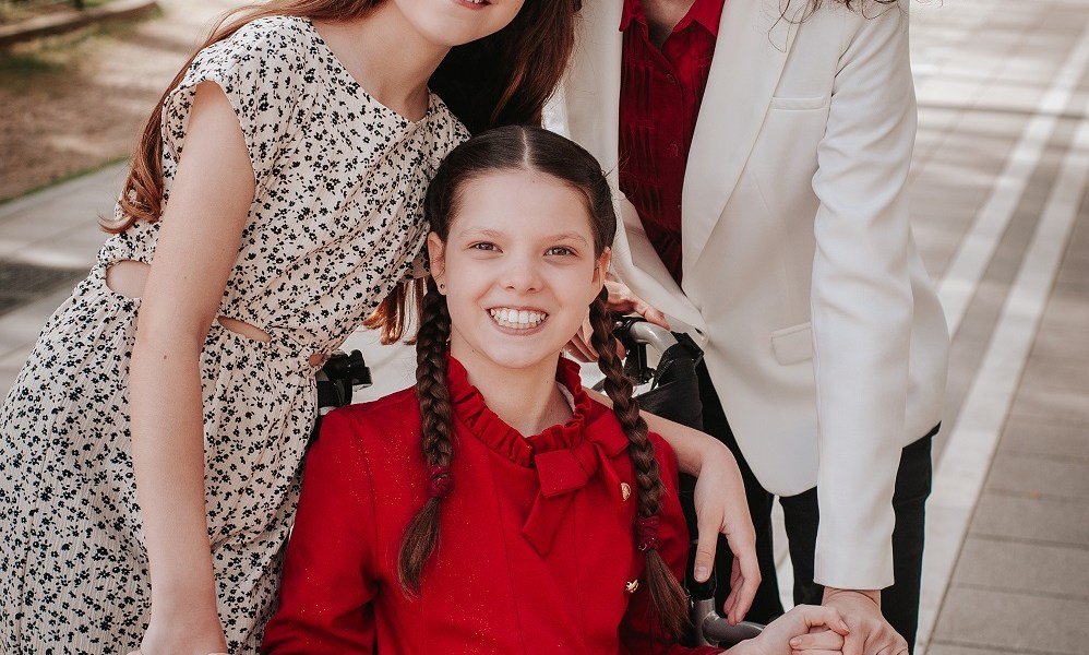 Carolina, in a red dress with braids, smiles in her wheelchair, embraced by her family, whose faces are cut off by the frame.