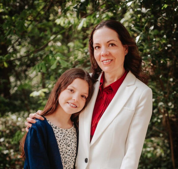 A long-haired girl in a navy cardigan poses in the arms of her mother, who wears a white jacket and red blouse, against a leafy background.