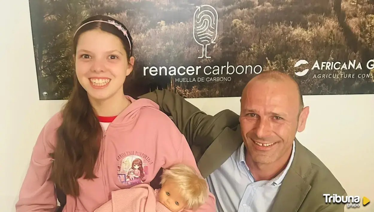 A girl in a pink Carolina Purpurina hoodie, holding a doll, poses with a man in front of the Renacer Carbono banner.